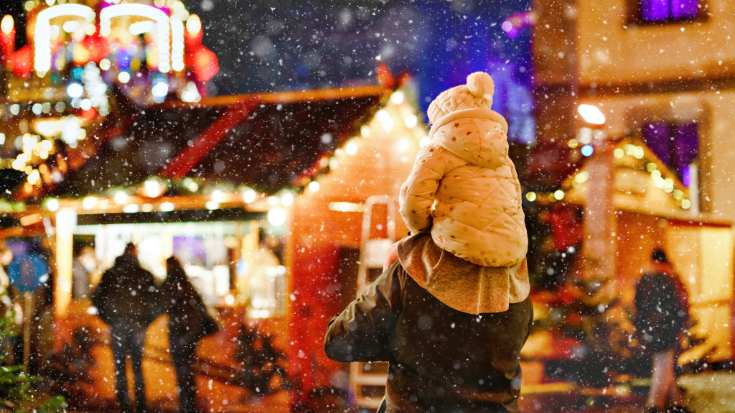 An image of a parent giving their small toddle-age child a piggy back from behind. It is night time and they are both wrapped up in heavy winter coats and hats and are walking around a brightly illuminated Christmas market as it starts to snow. To illustrate a blog post entitled 'Extreme Day Trips - UK Christmas Markets Edition'.