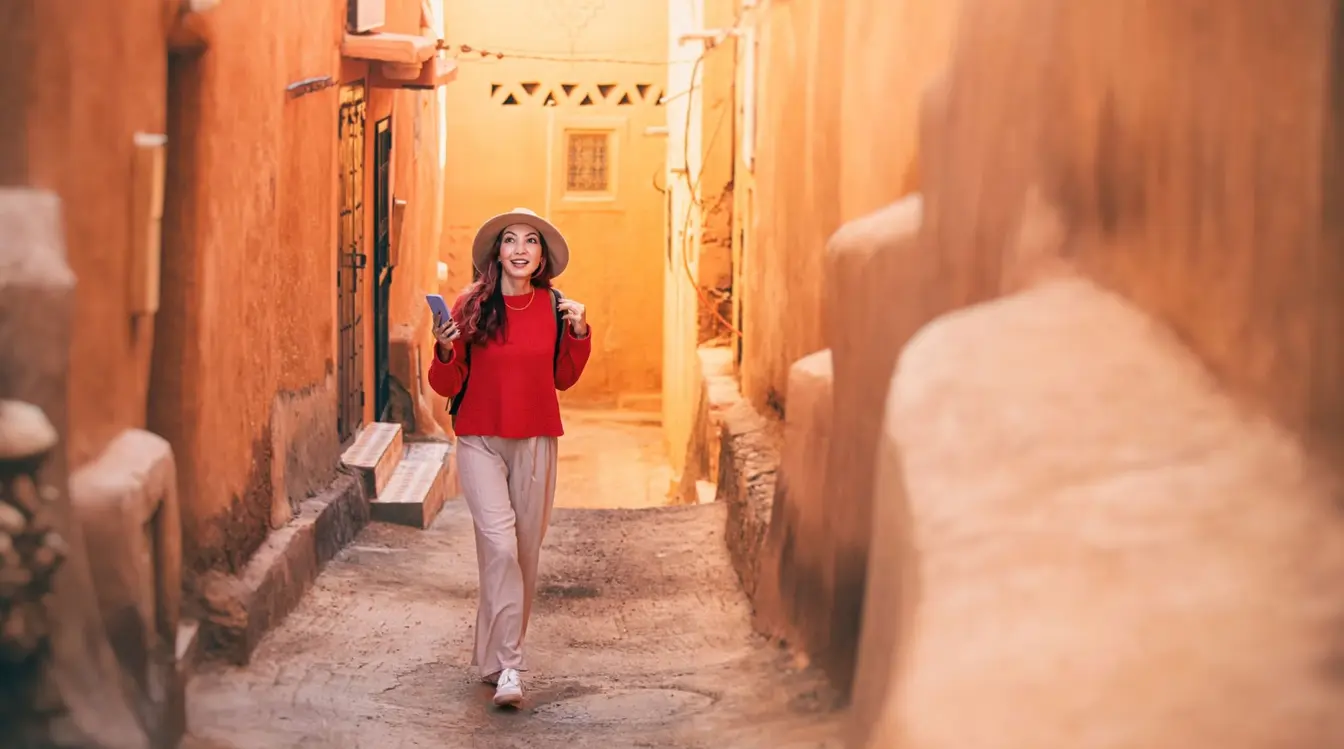 View of a woman holding a smartphone, walking through a clay-walled street in Ait Benhaddou, Morocco.