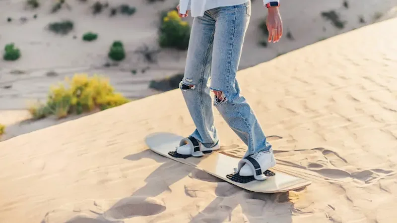 A woman's legs in blue jeans are attached to a sandboard, with sand dunes and desert plants visible in the background.