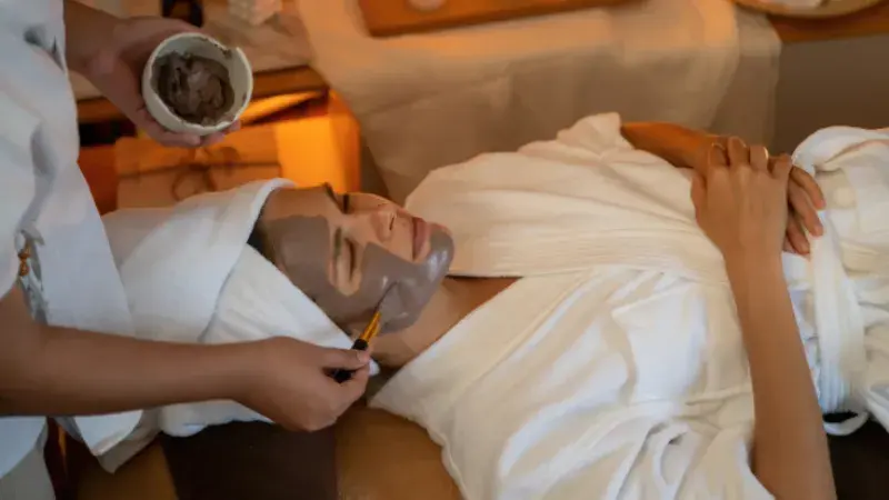A woman in a white bathrobe lying on a massage bed receiving a brown mud mask treatment at a beauty spa. 