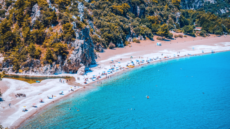 View of Cirali beach and Olimpos, Olympos mountain in a sunset light. Long curved bay on Turkey’s Antalya coast, with rows of beach umbrellas and swimmers enjoying the calm, bright blue sea. Behind the beach are forested hills and rocky cliffs — a popular hot holiday destination in October, perfect for travellers staying connected with eSIMs.