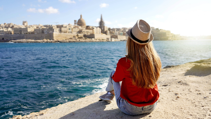 A photograph of a woman sitting on the edge of the sea looking out over a landscape of Valletta, Malta. You can see only her back and that she has long blonde hair, is wearing a straw sun hat wound with a black ribbon and she is wearing a red shirt and blue jeans. In the background, you can see the spires of the sandstone churches and buildings. To illustrate a blog post entitled 'Where is Hot in November in Europe?'