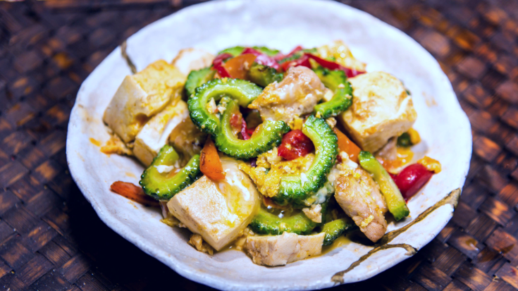 A colour photograph showing a close-up of one of Okinawa's most famous local dishes, goya chanpuru. On a bright white plate you can see cooked slices of the bitter melon plant goya, pork slices, egg, red peppers and tofu. To illustrate a blog post entitled 'Top Things to Do in Okinawa, Japan's Tropical Gem.'