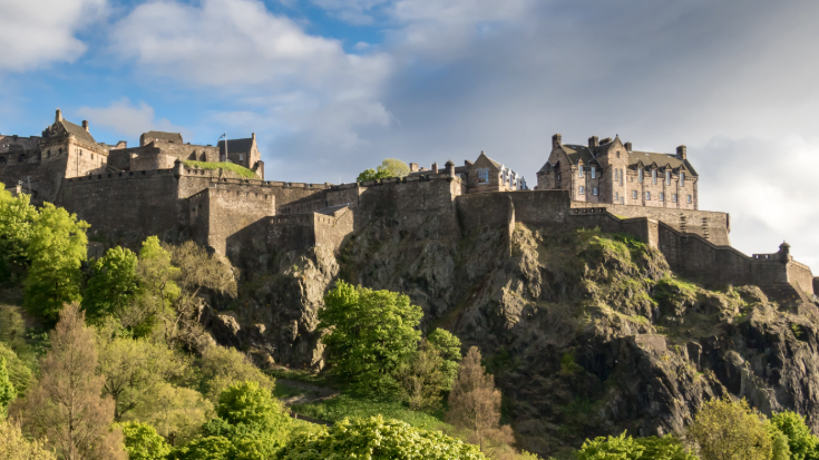 A colour photograph of Edinburgh Castle sitting on a red/brown cliff. It shows the building's walls and outhouses and underneath some greenery in the summer time, including trees and bushes. The sky in the background is a clear blue with some white clouds. To illustrate a blog post entitled 'Guillermo Del Toro's Frankenstein Filming Locations.'