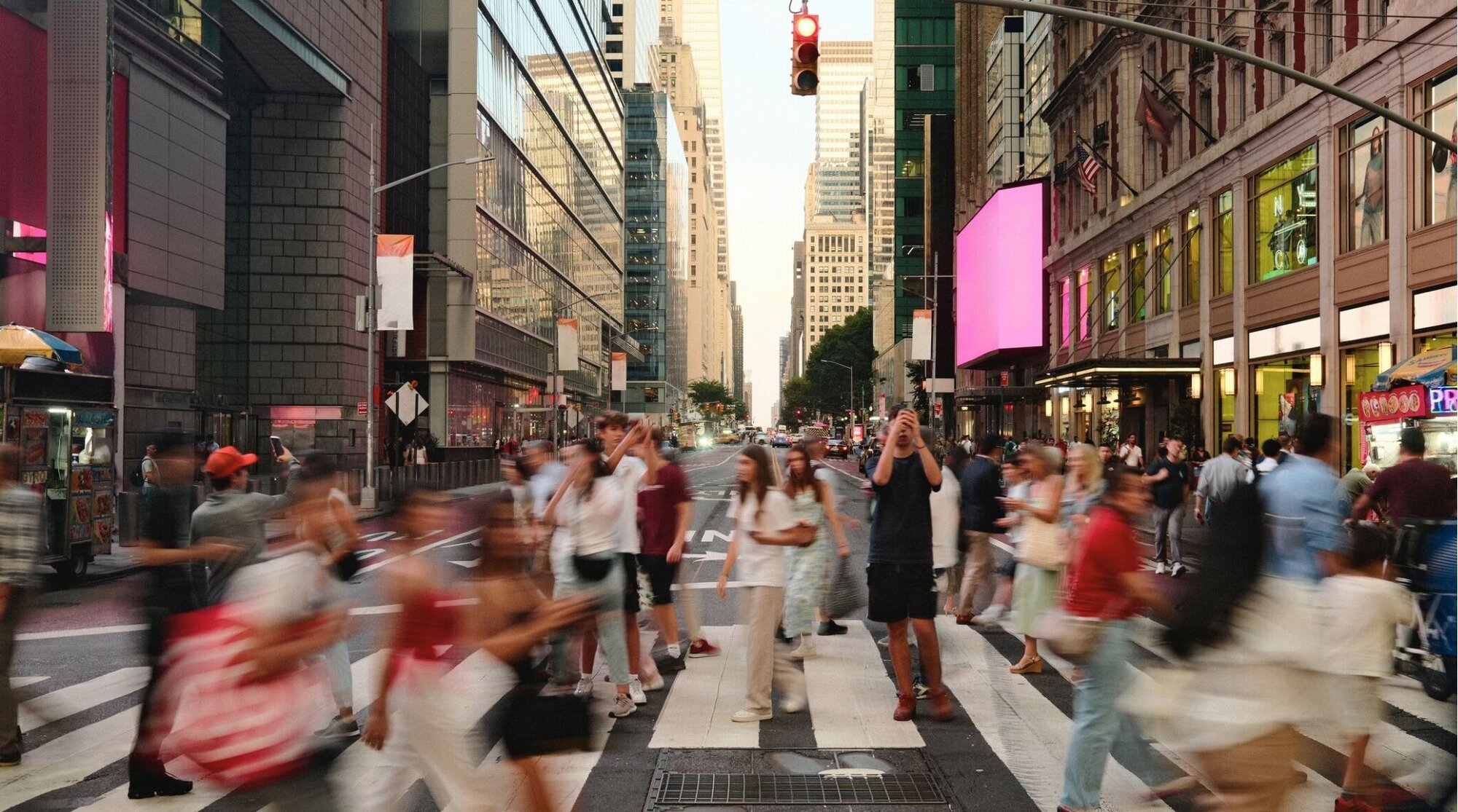 Multitud cruza un paso de peatones en una avenida de Nueva York, con movimiento y luces de ciudad.