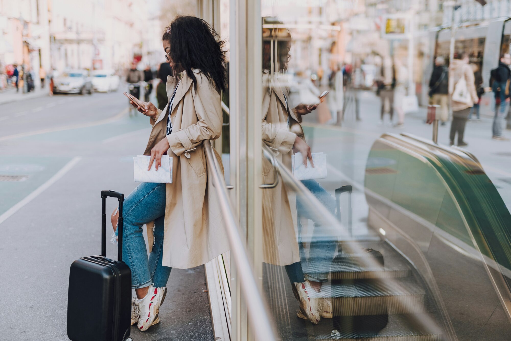 Woman with a suitcase and long coat, using her phone in a city