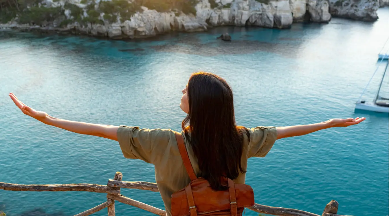 Turista de braços abertos contemplando a baía de Cala Macarella, em Menorca, com veleiro ao fundo nas águas turquesa das Ilhas Baleares.