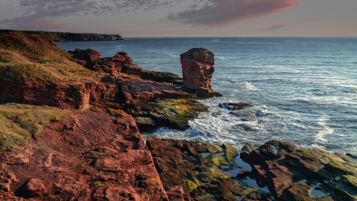 A color photograph showing the beautiful but choppy waters off the Scottish coast at Devil's Head in Arbroath, Scotland. It shows a towering red sandstone sea stack at Seaton Cliffs and colourful blue and pink sunrise. To illustrate a blog post entitled 'Guillermo Del Toro's Frankenstein Filming Locations.'