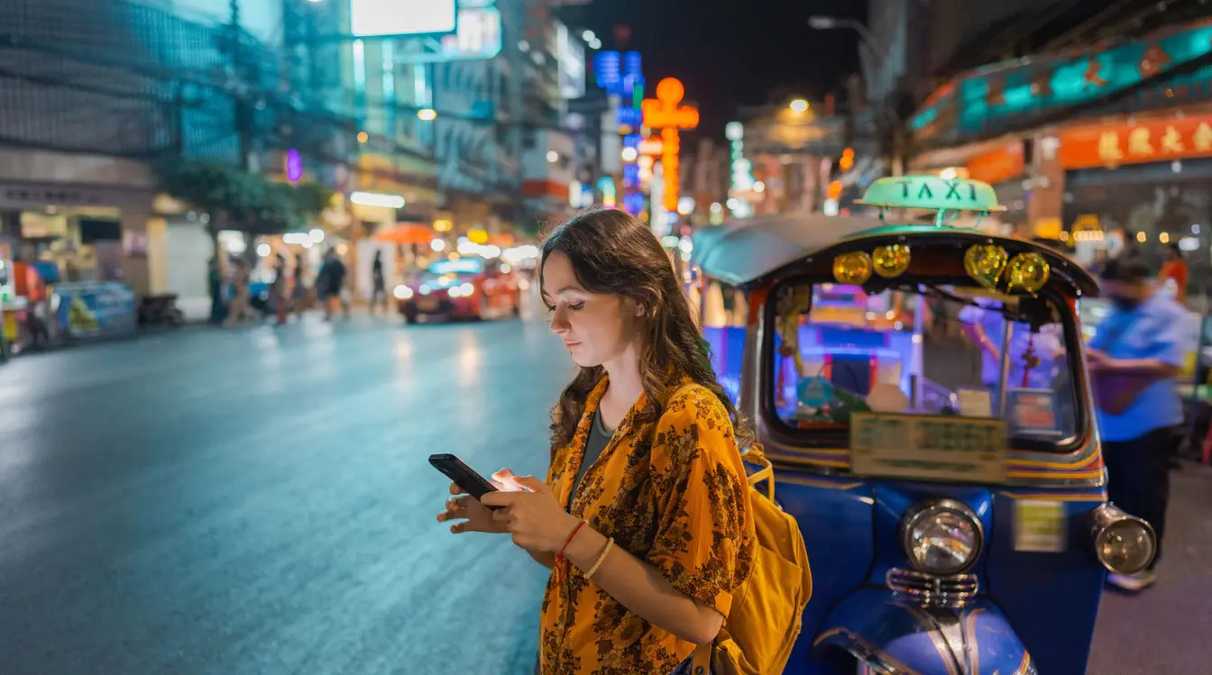 A young woman using a smartphone next to a tuk-tuk on a brightly-lit street at night in Chinatown, Bangkok.