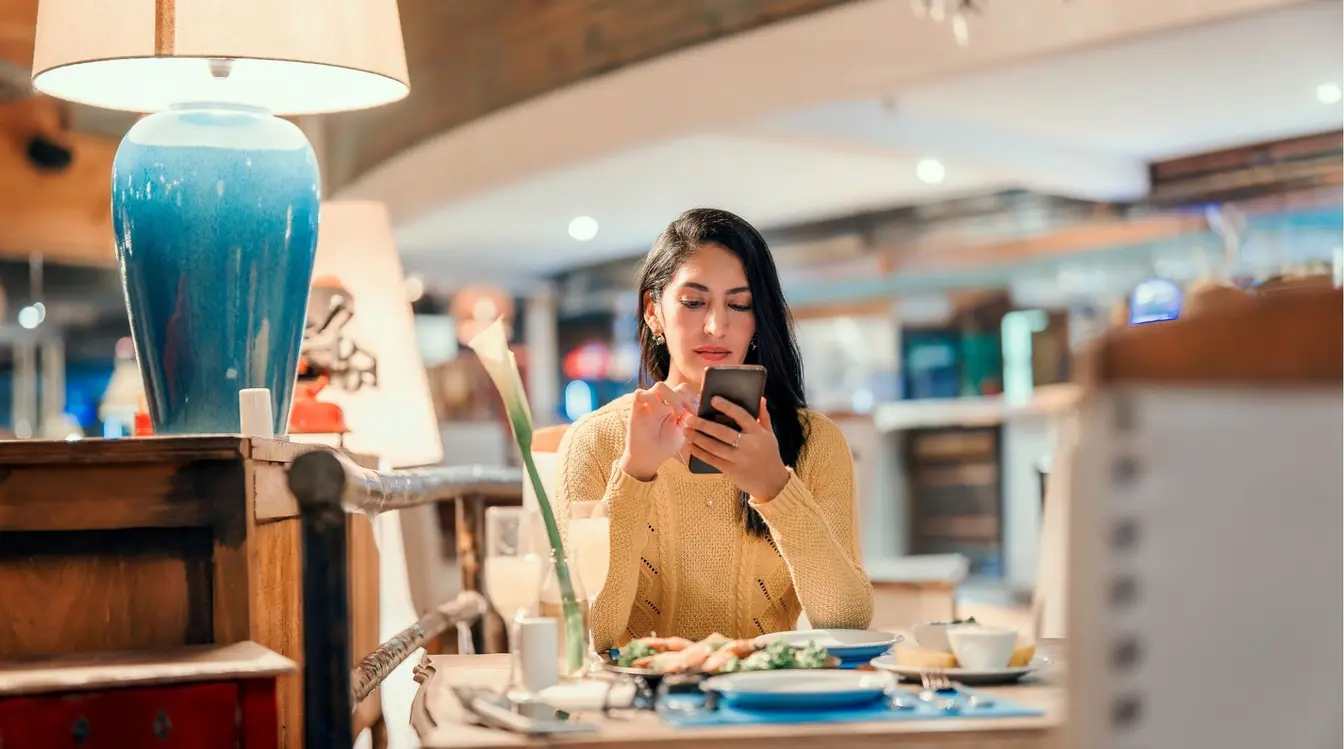 Mulher de cabelos escuros sentada à mesa de um restaurante, comendo uma salada em uma tigela branca enquanto olha para a tela do celular em suas mãos.