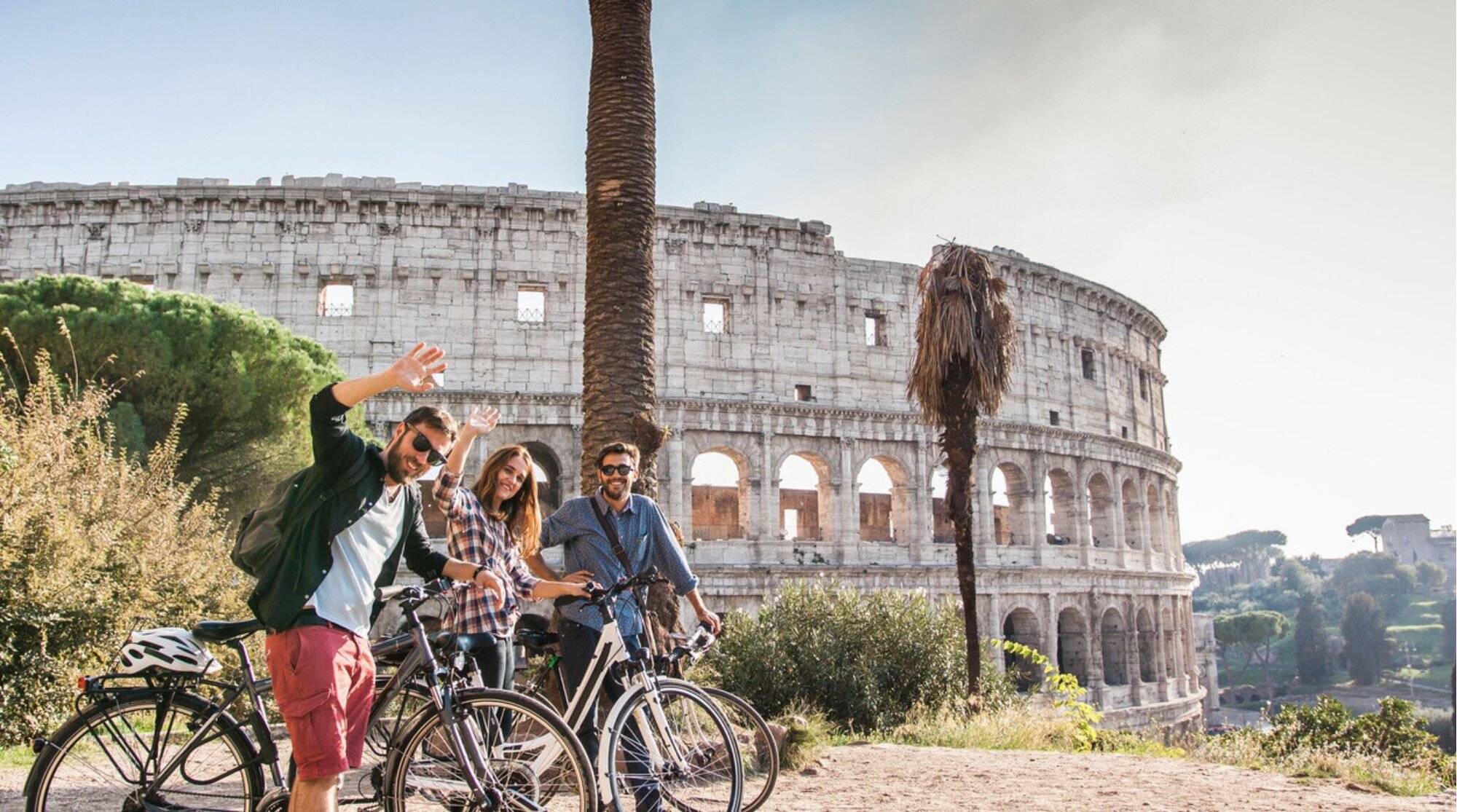 Três jovens amigos turistas felizes com bicicletas no Coliseu em Roma se divertindo posando e acenando para a câmera.