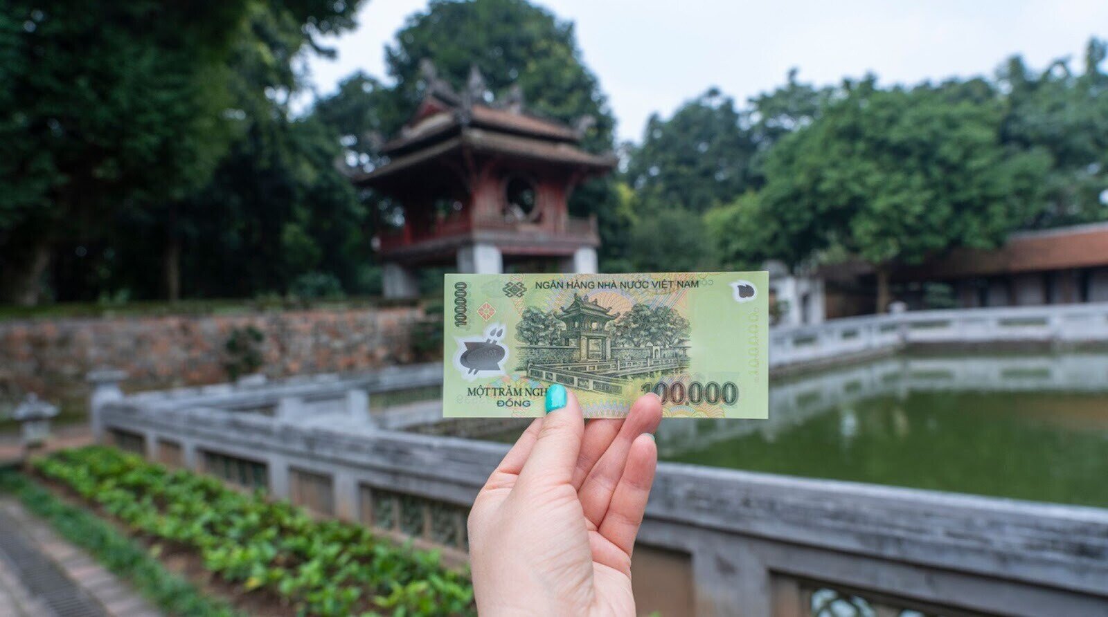 personne montrant un billet de 100 000 dongs vietnamiens devant un bassin et un temple religieux.