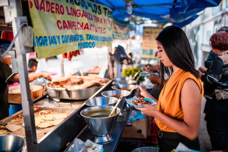 Mujer comprando comida en un puesto callejero en México, con ollas y guisos al fondo.
