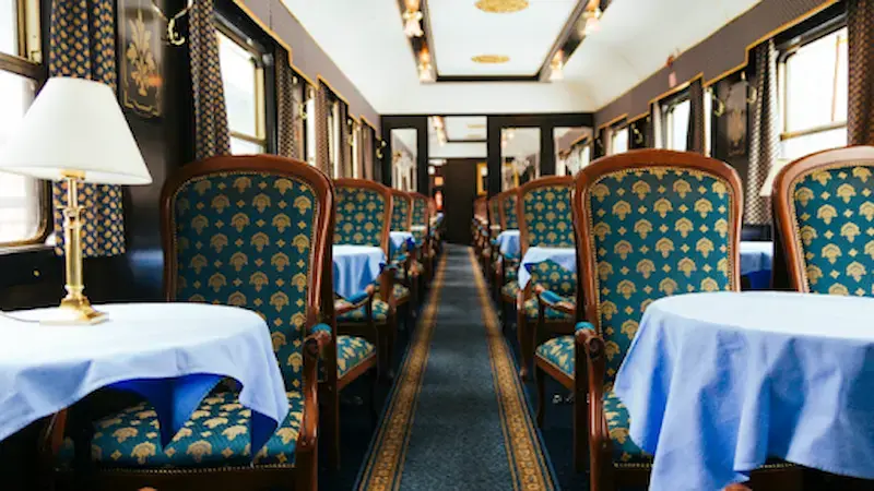 A luxury train carriage showing mahogany chairs with gold and blue cushions, dressed tables with gold lamps and blue carpets.  