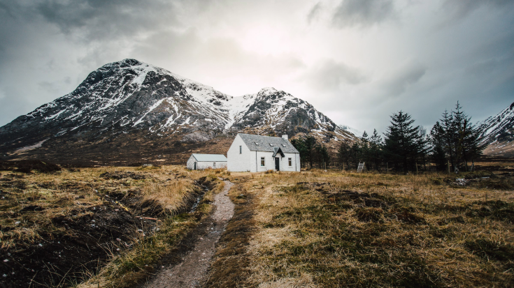 A colour photograph showing a desolate but beautiful whitewashed cottage in sitting in at the foot of a snow-capped peak. In the foreground is yellowing grass and a dirt path and in the background the sky is grey with dramatic grey clouds hanging overhead. The area is Glencoe in the Scottish Highlands. To illustrate a blog post entitled 'Guillermo Del Toro's Frankenstein Filming Locations.' 