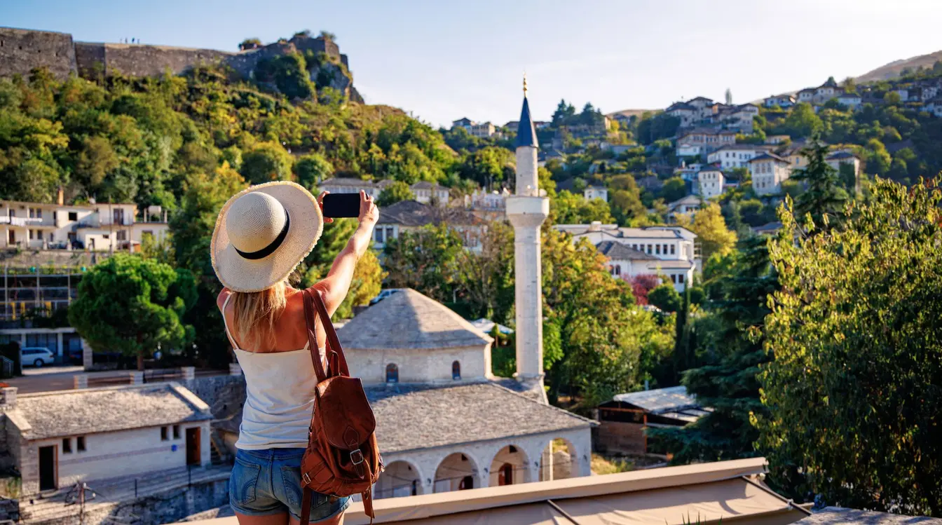 A woman in a straw hat takes a photo of a landscape in Gjirokaster, Albania, with green hills in the background.