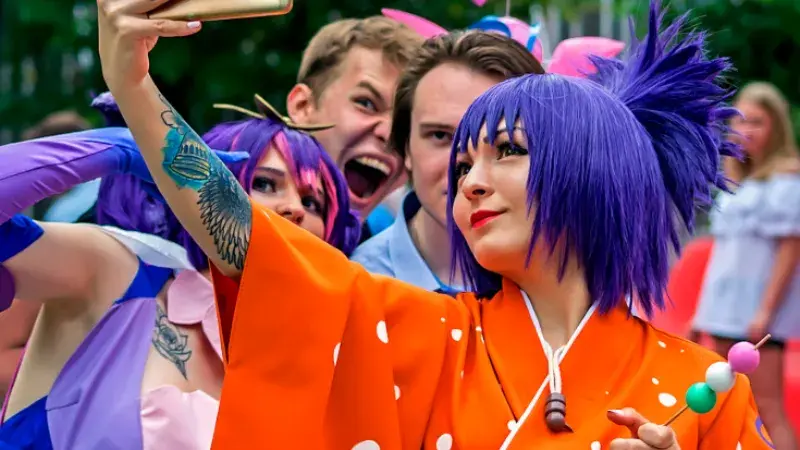 A cosplaying woman in a purple wig and an orange yukata, holding up her mobile phone to take a selfie. 