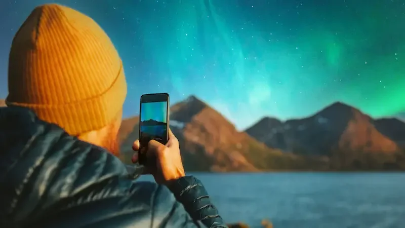 A man in an orange hat from behind as he takes a picture of the patterns of the Northern Lights in the Norwegian night sky.