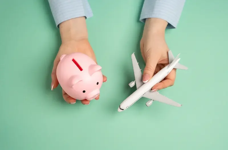 飛行機の模型とピンクの貯金箱を持つ手、旅費の節約や予約のイメージ。