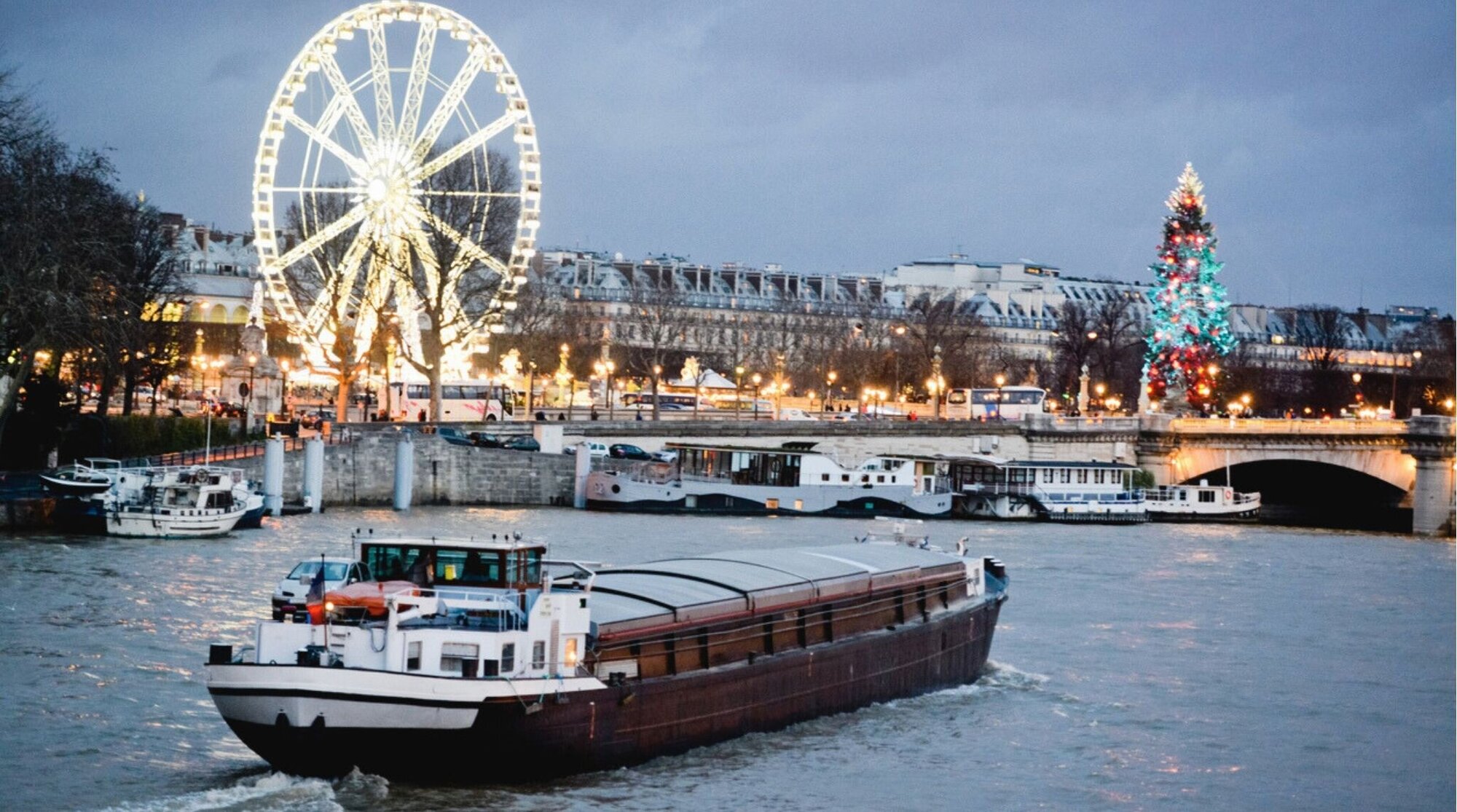 Barco navegando por el río Sena en París durante Navidad con luces festivas y ambiente invernal al anochecer.