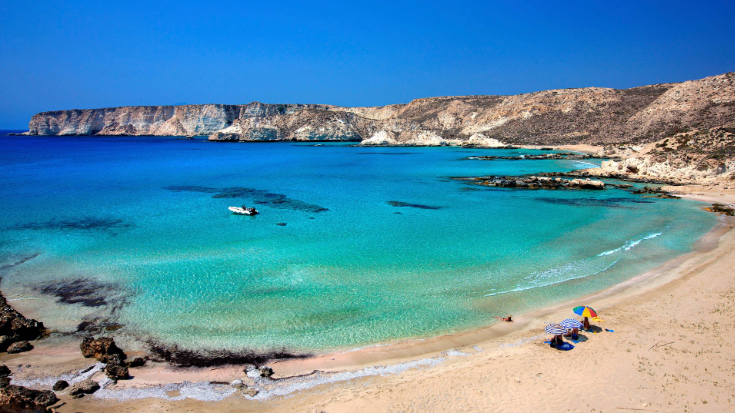 A photograph showing a landscape view of a beach in Koufonissi, a tiny uninhabited island, just south of Crete in Greece. The sands are white and the sea is a clear blue. On the beach are two parasols, one red and one with blue and white stripes. The sky is clear and bright blue and the sunshine is out. To illustrate a blog post entitled 'Where is Hot in November in Europe?'