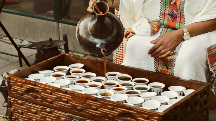 This is a photograph showing a close-up on an Ethiopian woman's hands as she pours black coffee from a large traditional ethiopican coffee jog into many small white empty coffee cups sitting together in a large dark straw basket. It is an example of how a traditional Ethiopian coffee ceremony may look. To illustrate a blog post entitled 'Which Country Has the Best Coffee Culture?'