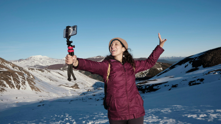 A photograph of a woman wearing a maroon/deep red-coloured 3/4-length puffer coat, a beige knitted hat and long dark brown hair. She is holding a camera tripod, which has a camera attached to it, and is taking a selfie with her arms outstretched and her mouth open in a happy expression. In the background is a set of snowy mountains and a clear blue sky. This is an image to illustrate a blog post entitled 'Unlimited eSIM: Plans, Benefits, and How It Works'. 