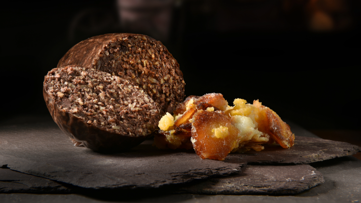 A close-up full colour image of a round of haggis, which is many shades of brown with white mottles of animal fat throughout. The haggis has been cut open with one slice laid out before it on a wooden board, ready to be served to guests for Burns Night. To illustrate a blog post entitled 'What is Burns Night and How to Celebrate It'.