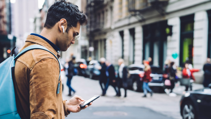 A photograph of a young man standing on the street in New York City, with some sandstone buildings next to him. He is looking down at his phone while wearing in-ear headphones.