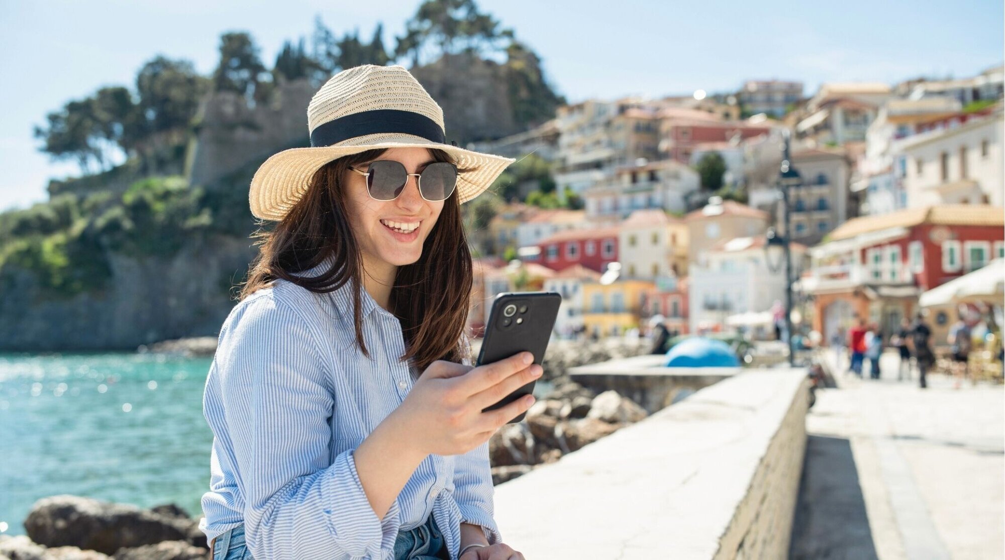 Mujer con sombrero y gafas usando el móvil junto al mar, pueblo costero de fondo en un día soleado.