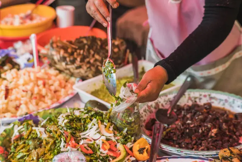 Manos preparando ingredientes para tacos en un puesto de comida, con salsas y guarniciones sobre la mesa.