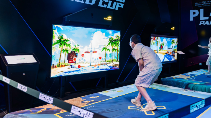 Gaming enthusiasts playing a ball-throwing game in front of a big screen on a padded surface at the Esports World Cup festival area.