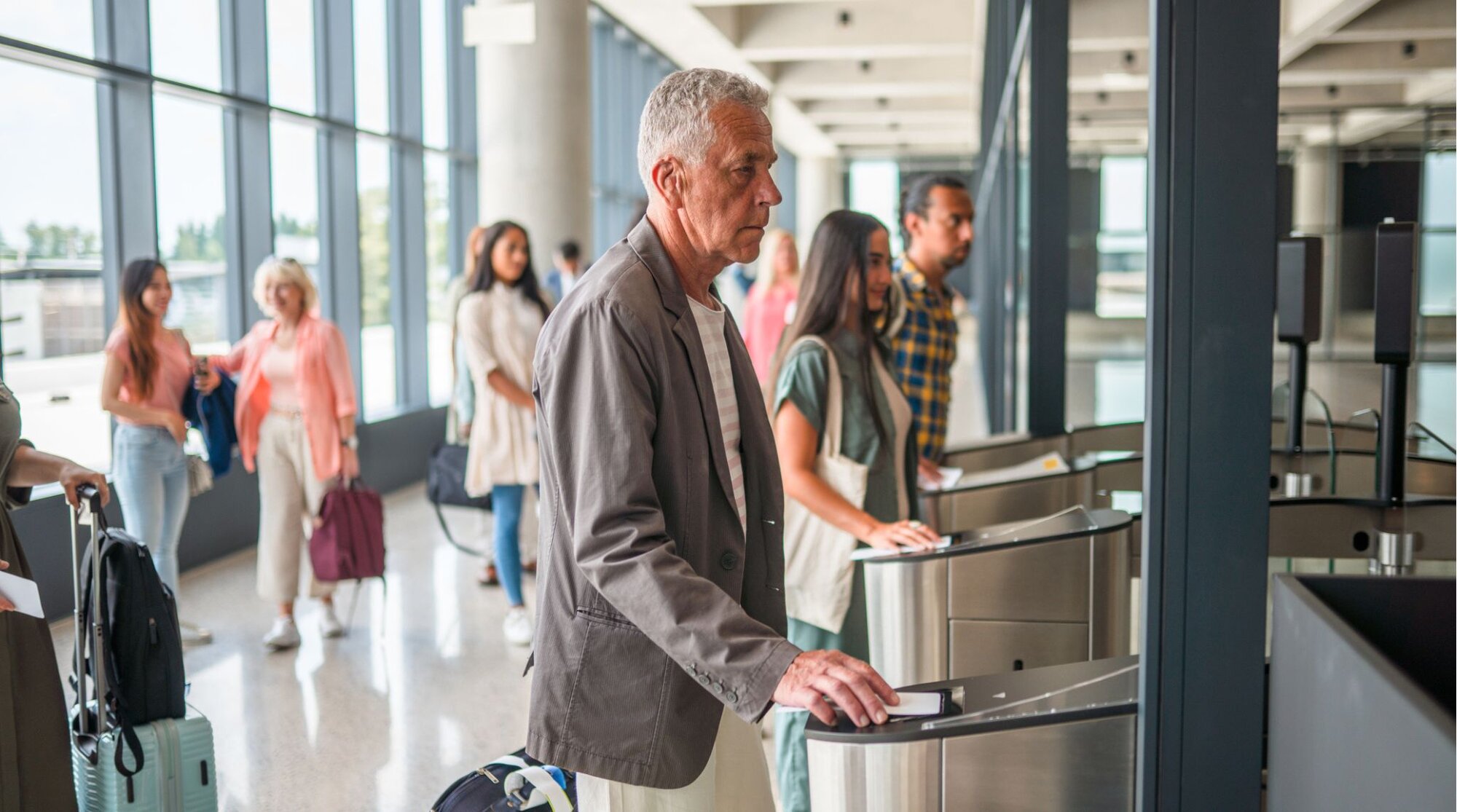 Viajantes passando por totem de controle automatizado em aeroporto, com fila de passageiros ao fundo.