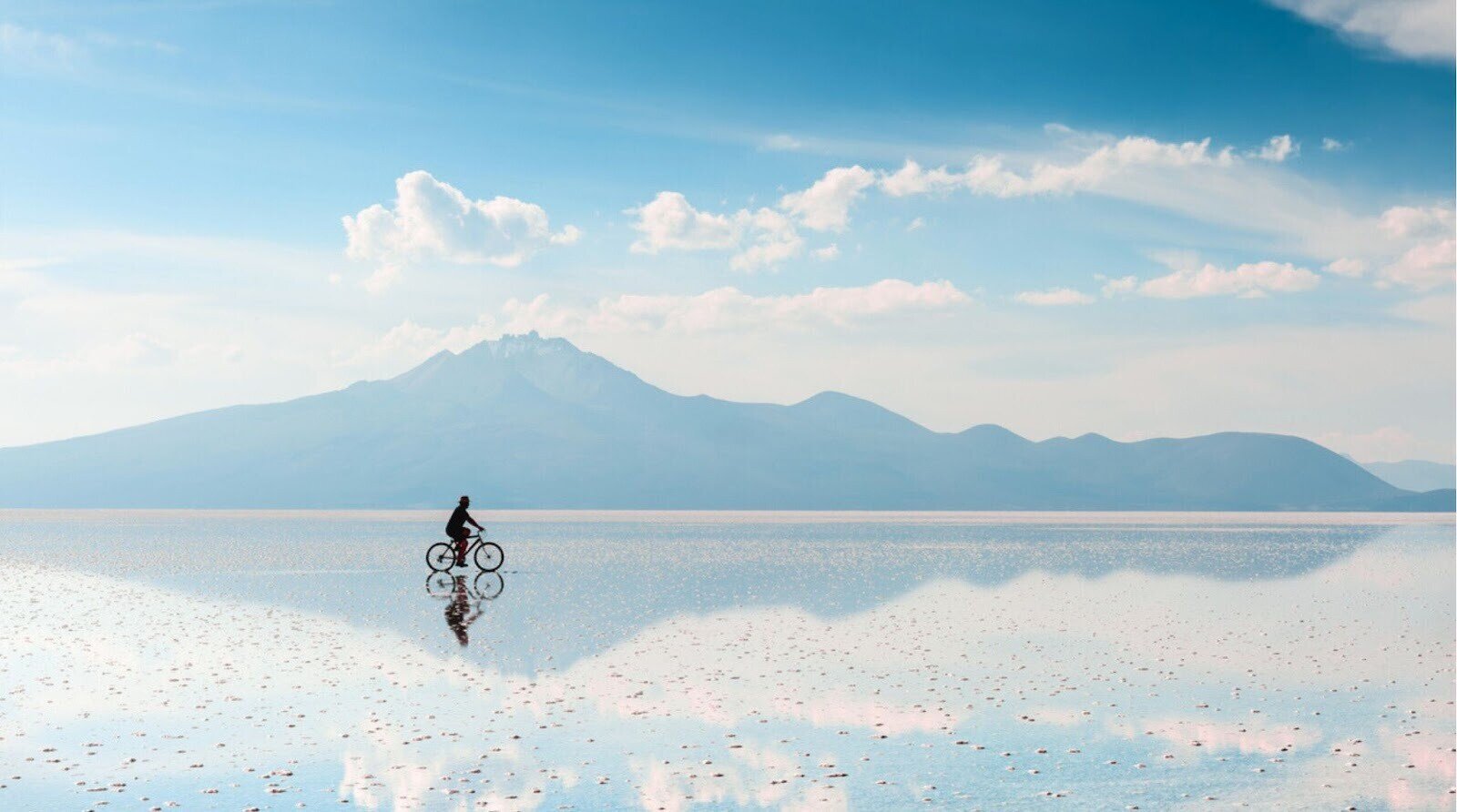 Turista anda de bicicleta no Salar de Uyuni, na Bolívia. As montanhas e o céu se refletem na superfície da água.
