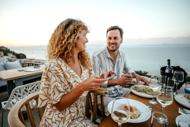 Mujer tomando una foto de su comida en una terraza frente al mar durante una cena.