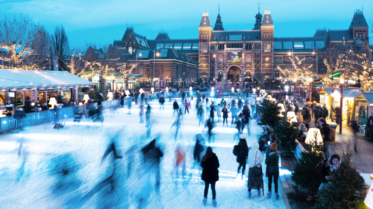 A colour image that shows a large rectangular ice rink in the foreground, with skaters dressed in winter clothes whizzing across it. In the background is Amsterdam's famous Rijksmuseum building and around the edges of the rink are small festive huts for the city's Christmas market.To illustrate a blog post entitled 'Extreme Day Trips: Eurostar Christmas Markets Edition'.