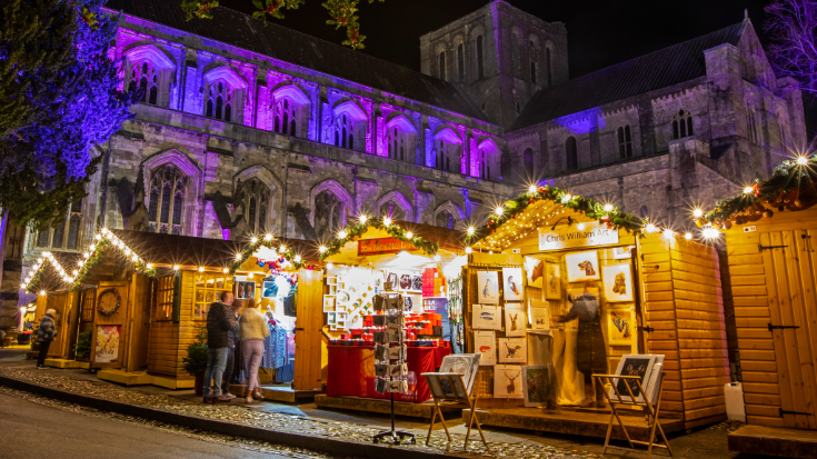 A view of the Christmas Market in the grounds of the historic Winchester Cathedral in the city of Winchester, Hampshire, UK. In the background is the black night sky and Winchester cathedral, which is low and the longest Medieval example in the world. In the foreground you can see vendors selling Christmas goods and artwork from traditional german-style wooden huts. To illustrate a blog post entitled 'Extreme Day Trips - UK Christmas Markets Edition'. 