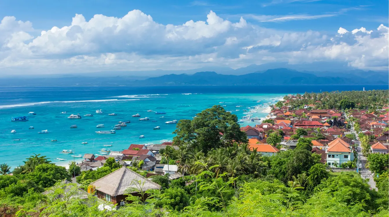 View of Nusa Lembongan's coast, with houses between the trees, boats on the water, and mainland Bali in the background.