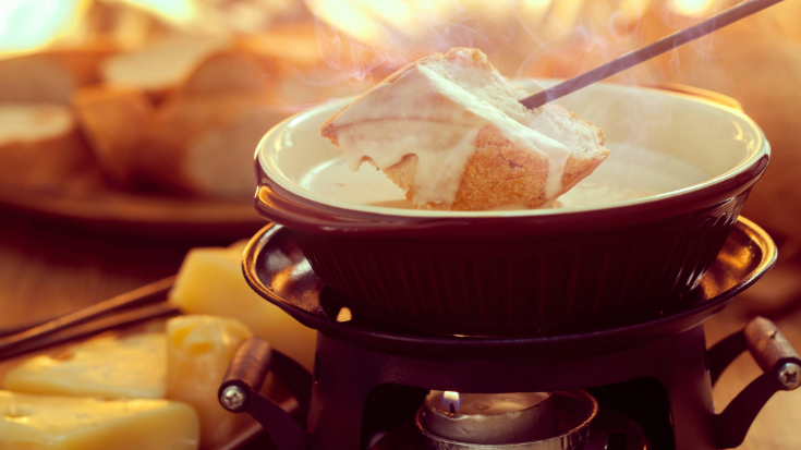 A close-up photograph of cheese melting over a square of bread, suspended over a fondue bowl, showing the idea of eating a cheese fondue at a specialist cheese bar in Camden, to illustrate a blog entitled 'Things to do in London in December'.