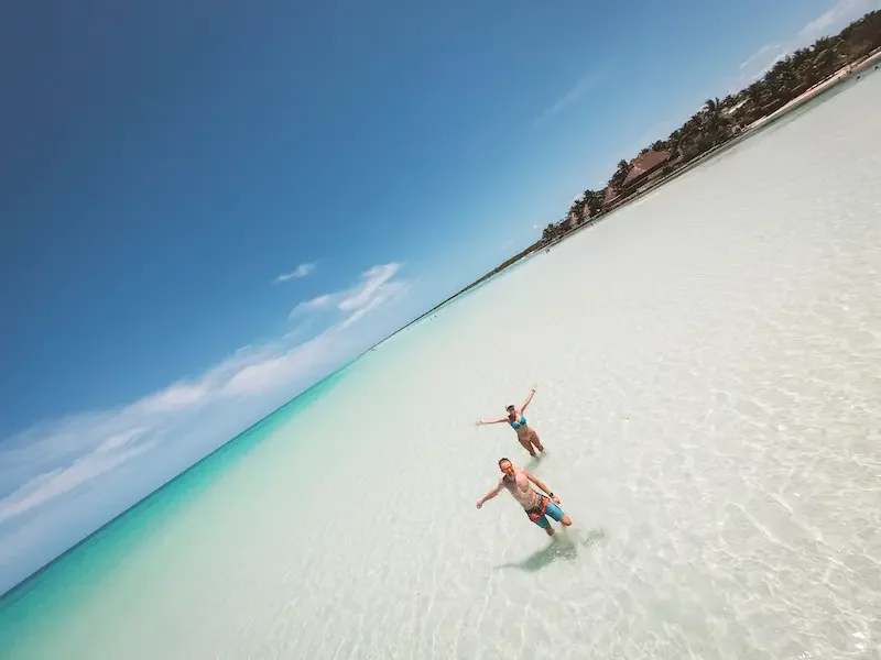 Pareja caminando por aguas poco profundas en Isla Holbox, con mar turquesa y playa de arena clara.