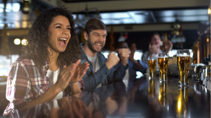 A young woman in a flannel shirt sits at the bar watching a sports game and cheering, next to a man with a beard who is also watching the 2027 Six Nations games.