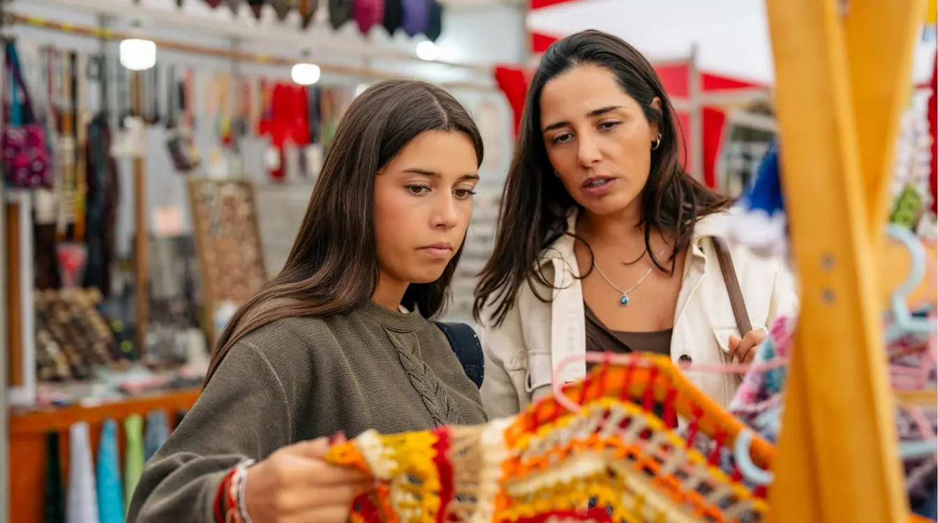 Duas mulheres jovens em uma feira de artesanato olhando atentamente para peças de tecidos andinos coloridos.