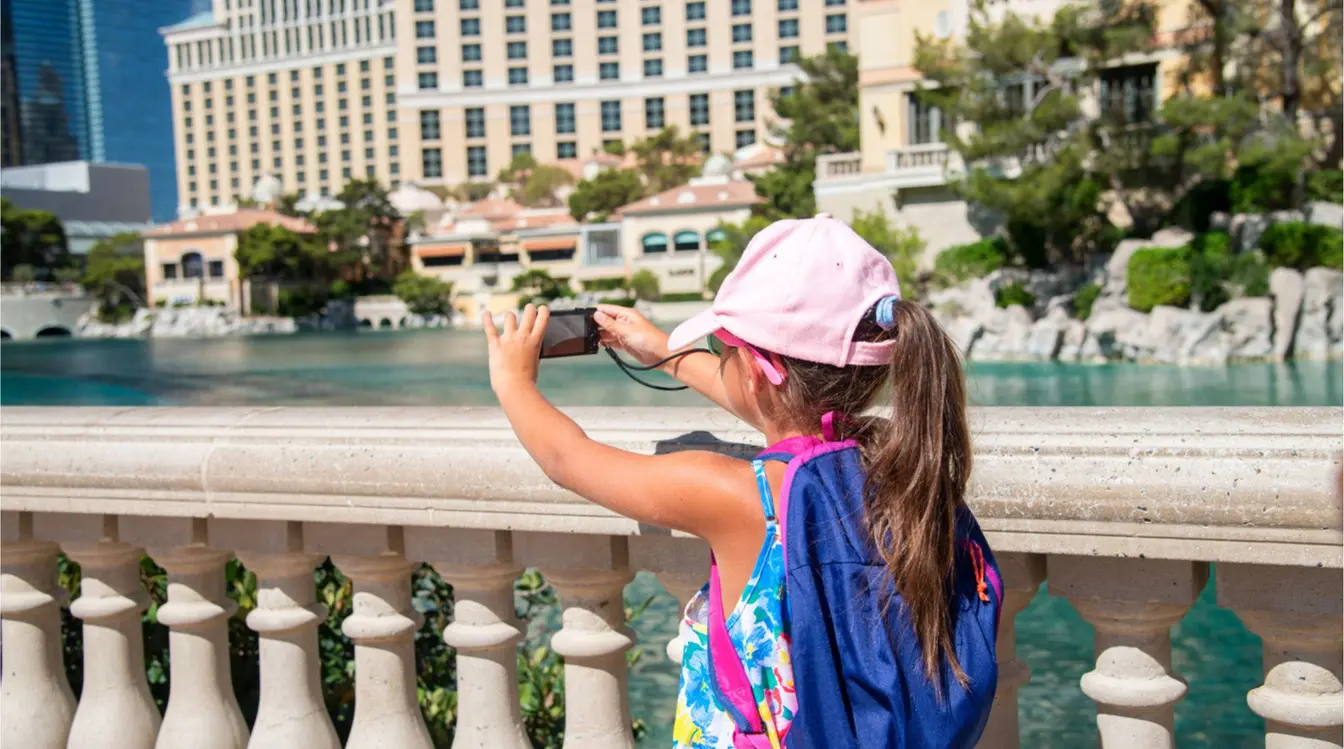 Niña con gorra rosa tomando una foto frente al lago del hotel Bellagio en Las Vegas, con edificios del Strip al fondo en un día soleado.
