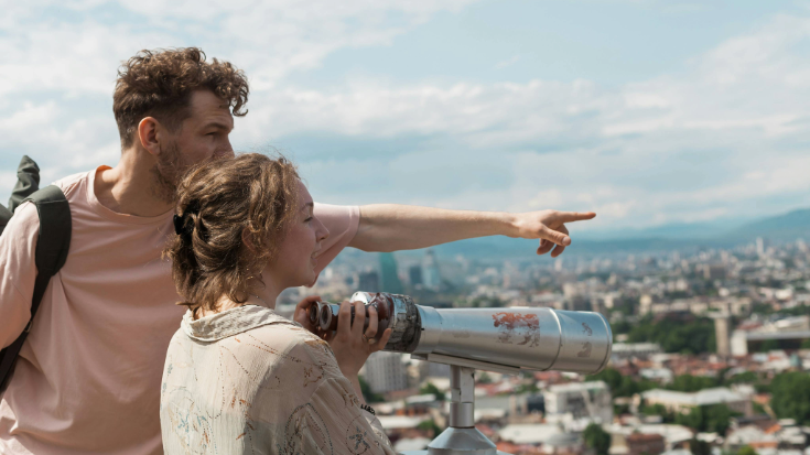 A photograph with a couple in the foreground. He is wearing a pink t-shirt, a back pack and has brown hair and is pointing out over a view. She is looking through a telescope and appears to be paying attention to whatever he is saying or pointing at. She also has brown hair, is wearing it tied up and has a cream-coloured shirt on. In the distance is a beautiful view of a city with hills and buildings all around. This is an image to illustrate a blog post entitled 'Unlimited eSIM: Plans, Benefits, and How It Works'. 