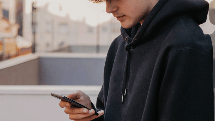 A young man wearing. black hoodie looking down at his mobile phone, with a city in the background. 