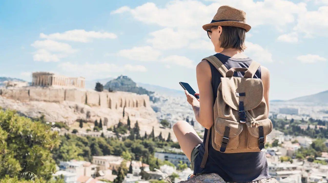 A solo female traveller sat holding her phone with a view of the Acropolis in Athens.