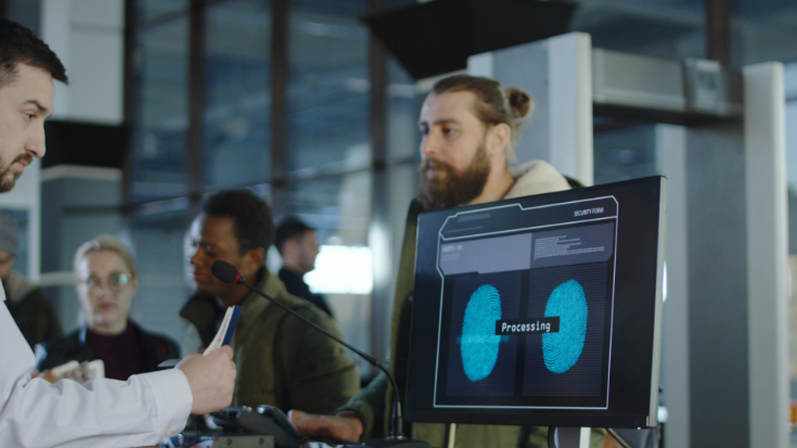 A photograph set in an airport, showing an airport customs officer looking at a computer screen. On the screen are two blue images of fingerprints againsta. black background. across the screen it says 'Processing'. In the background is a man with a beard and brown hair and some other travellers, waiting for their biometric data to be processed, as part of the EES system. To illustrate a blog post entitled 'Will ETIAS and EES Delay You? What UK Travellers Need to Know'?