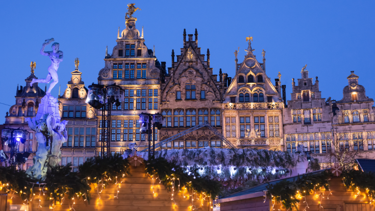 A colour image of a row of very ornate traditional Belgian buildings, with a darkening sky in the background and a row of Christmas market stalls covered in red and gold decorations in the foreground. To illustrate a blog post entitled 'Extreme Day Trips: Eurostar Christmas Markets Edition'.