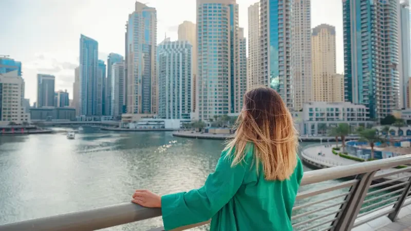 A woman with blonde hair in a green coat looks out over skyscrapers as she stands on the Dubai Marina Walk.