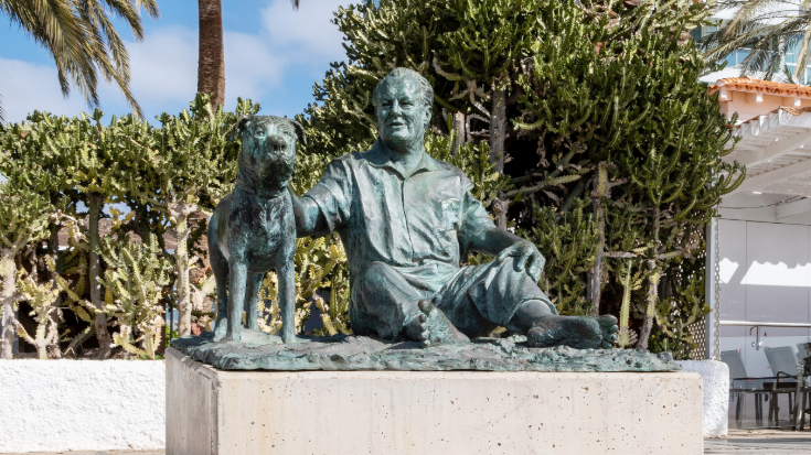 A life-sized bronze statue of former German Chancellor Willy Brandt sitting barefoot beside his faithful English basset hound, Bastian, in Morro Jable, Fuerteventura. The pair look relaxed under palm trees, celebrating the bond between people and dogs.